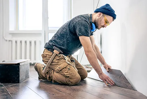 Worker kneeling on floor installing laminate flooring panels in a bright room with window light.