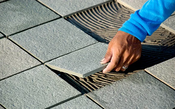 Close-up of a worker's hand placing gray ceramic tiles outdoors near a drain on a paved surface.