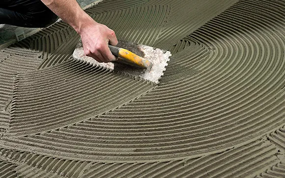 Worker smoothing gray ceramic tile adhesive with a notched trowel during flooring installation.