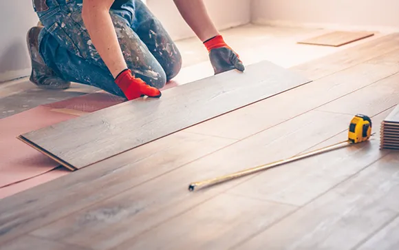 Close-up of worker's hands installing hardwood flooring planks with measuring tape on side.