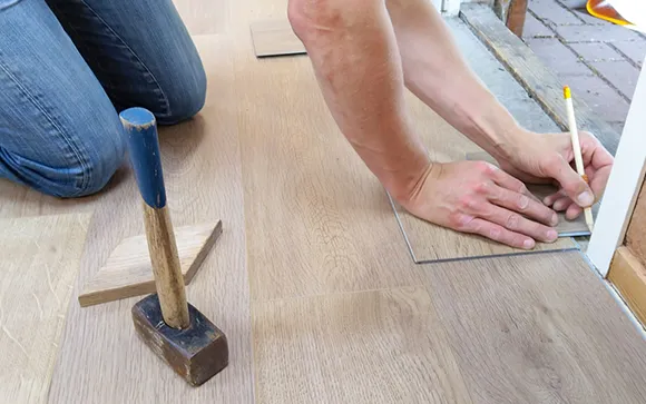 Close-up of hands using a mallet to install luxury vinyl plank flooring on a wooden subfloor.