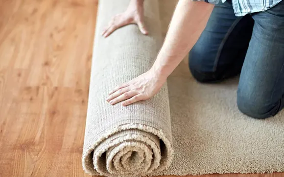 Person rolling up a beige carpet on a hardwood floor during carpet flooring installation.
